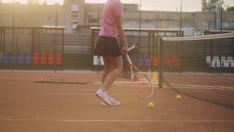 A-young-girl-is-tired-after-an-intense-outdoor-tennis-training.-Rolls-tennis-balls-along-the-net.